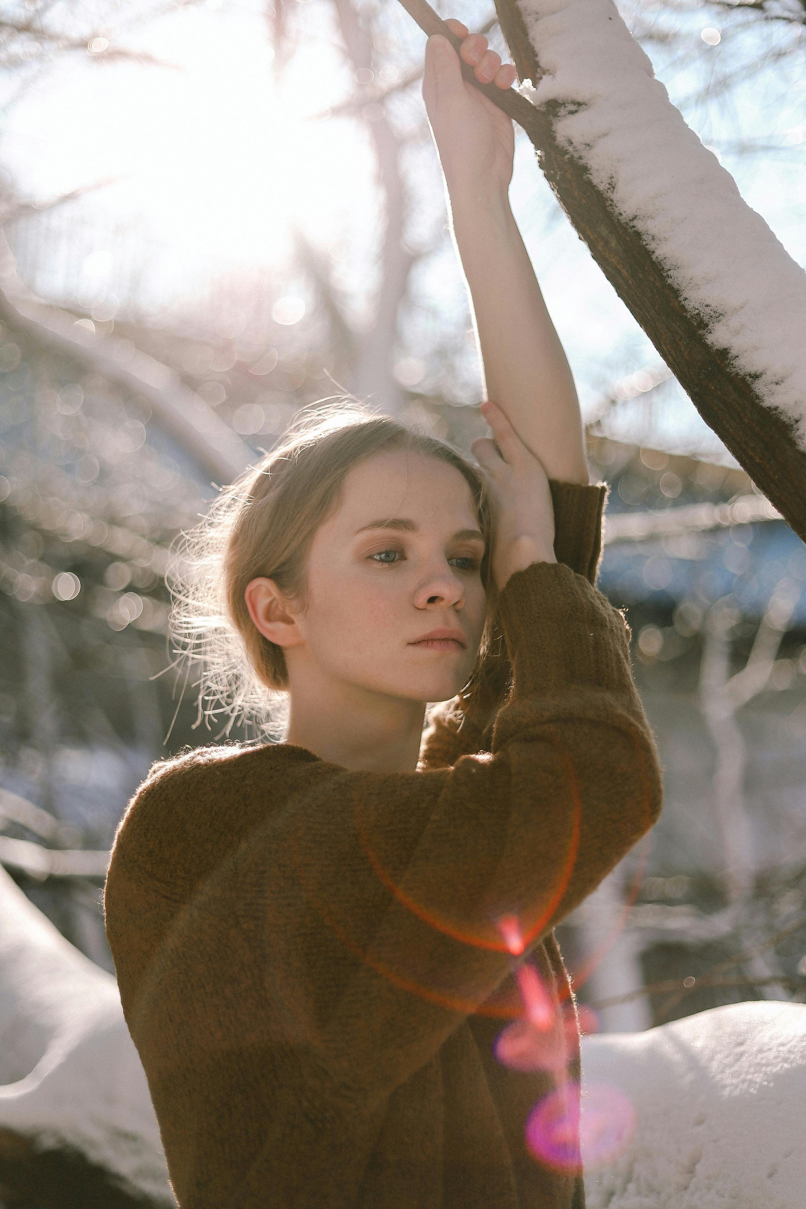 A female therapy client for anxiety and perfectionism stands alone in the winter, looking
somber. She is wearing a red sweater and is touching a tree.