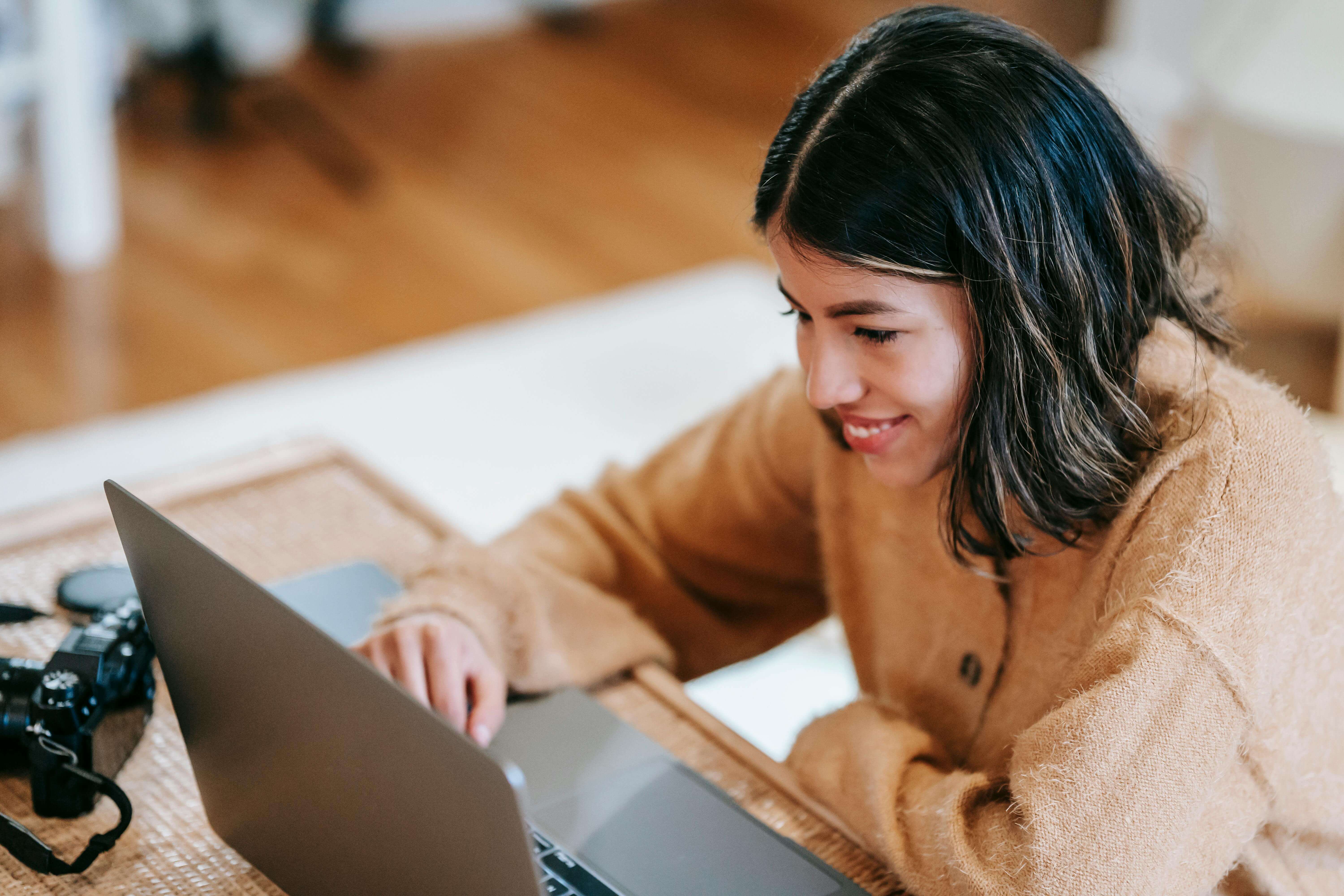 A brunette girl wearing a camel sweater is smiling at her laptop. She appears engaged
and happy following a perfectionism therapy telehealth appointment.